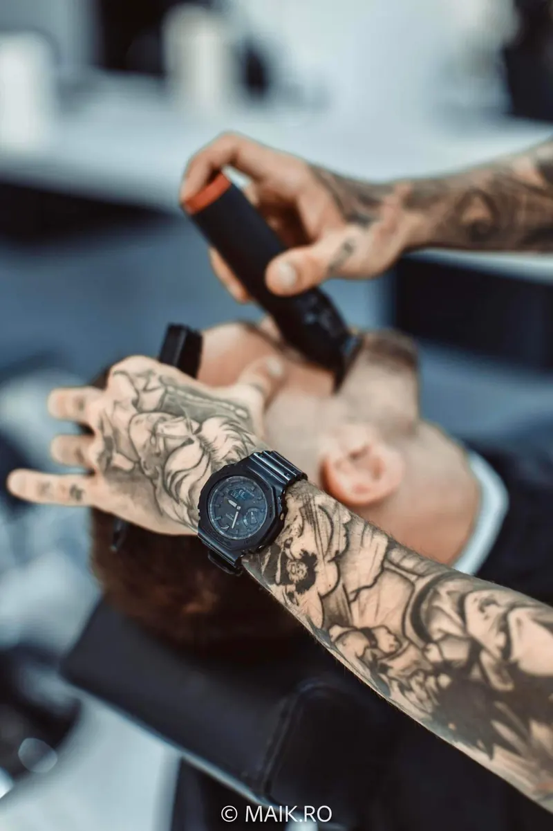 Frizer cu brațe tatuate și ceas negru, conturând cu mașina ceafa unui client.
