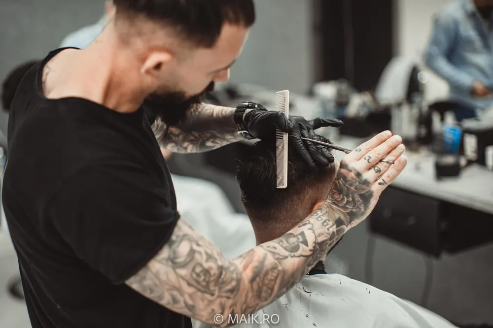 Frizerul, cu brațe tatuate și mănuși negre, tunde cu foarfeca peste pieptene părul unui client așezat pe scaun.